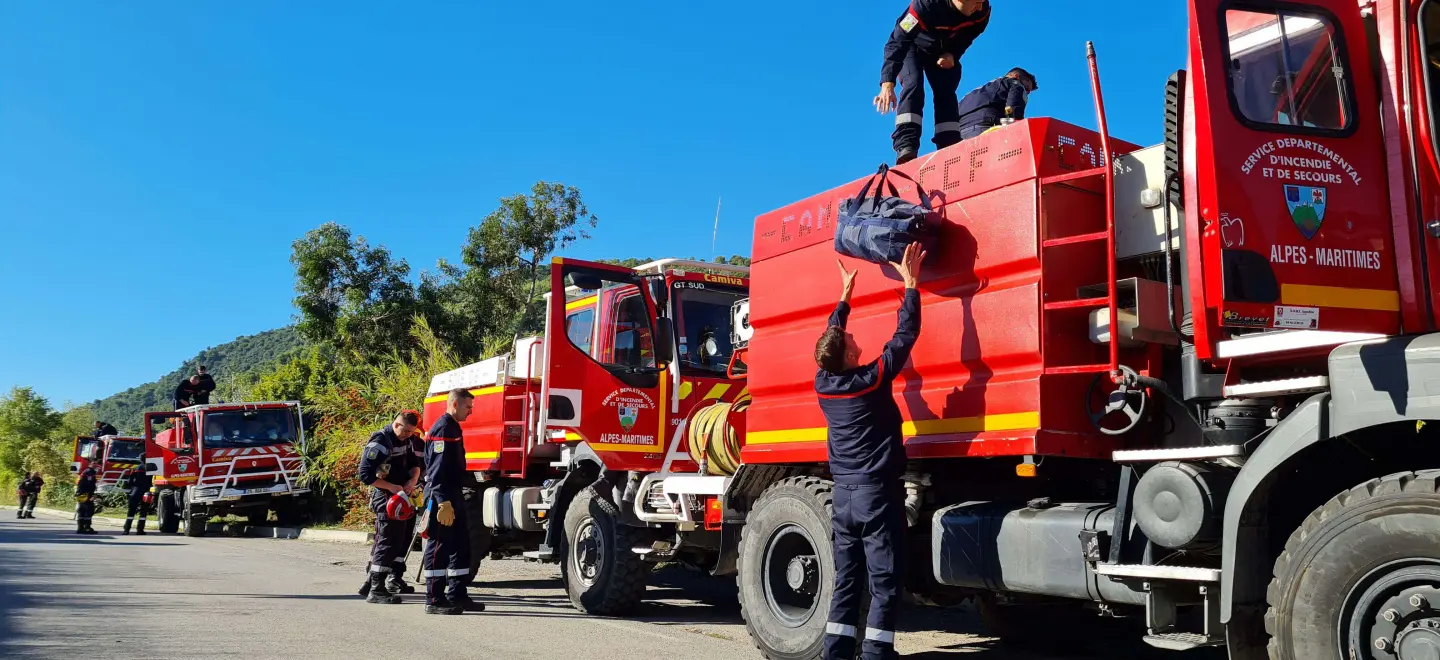 Des sapeurs-pompiers en uniforme s'activent autour de plusieurs camions rouges, identifiés comme appartenant au "SERVICE DÉPARTEMENTAL D'INCENDIE ET DE SECOURS ALPES-MARITIMES". L'un d'eux tend un sac à un collègue sur le toit d'un camion, sous un ciel bleu dégagé, dans un environnement montagneux.