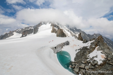 Petit lac de fonte glaciaire aux eaux intenses, de couleur cyan, niché dans un paysage montagneux. La neige recouvre les pentes environnantes, contrastant avec les affleurements rocheux et les sommets lointains.