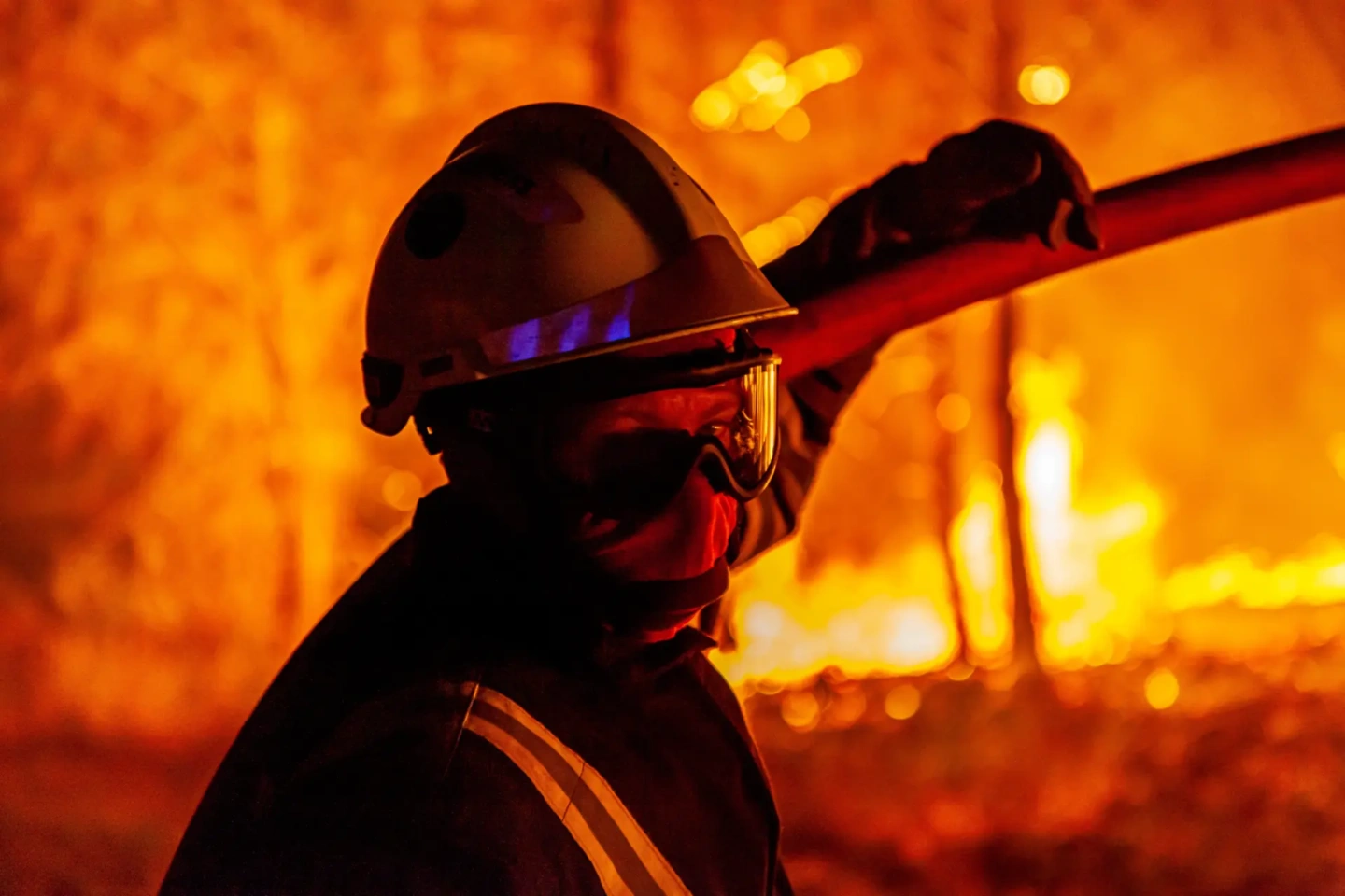 Un pompier est partiellement visible, en contre-jour devant les flammes intenses d'un incendie, tenant une lance d'incendie. Il porte un casque et des lunettes de protection.